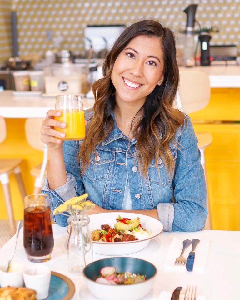 A young woman with dark hair at a table with breakfast and orange juice in hand at Farmhouse Corner Market in San Luis Obispo