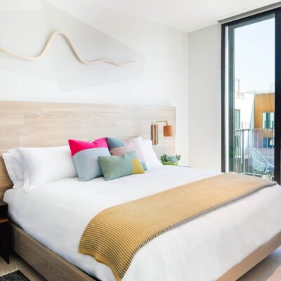 A clean bed with white sheets and colorful pillows, with a view out of a sliding glass door of the Hotel San Luis Obispo room's balcony.