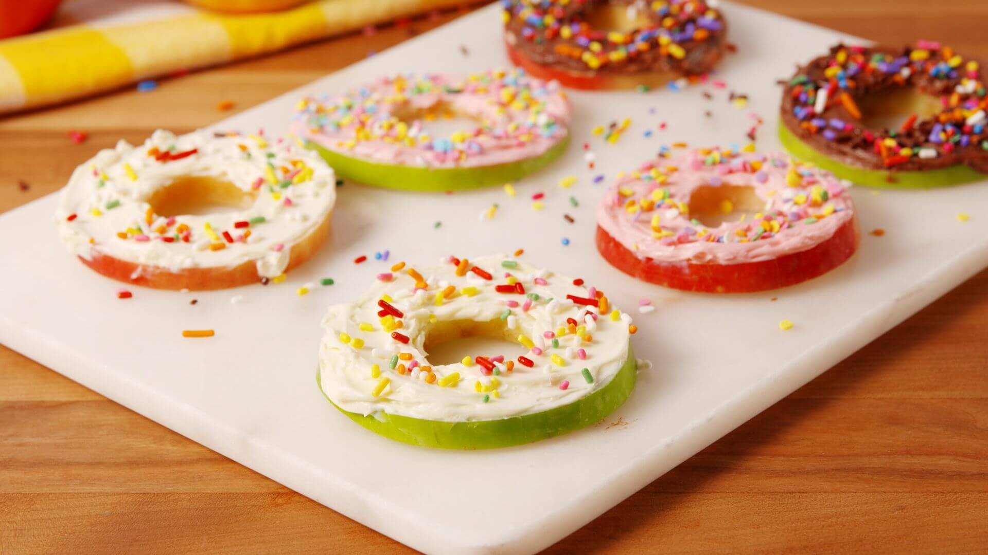 Six ring-shaped apple slices with thin layers of frosting spread over them, topped with colorful sprinkles.