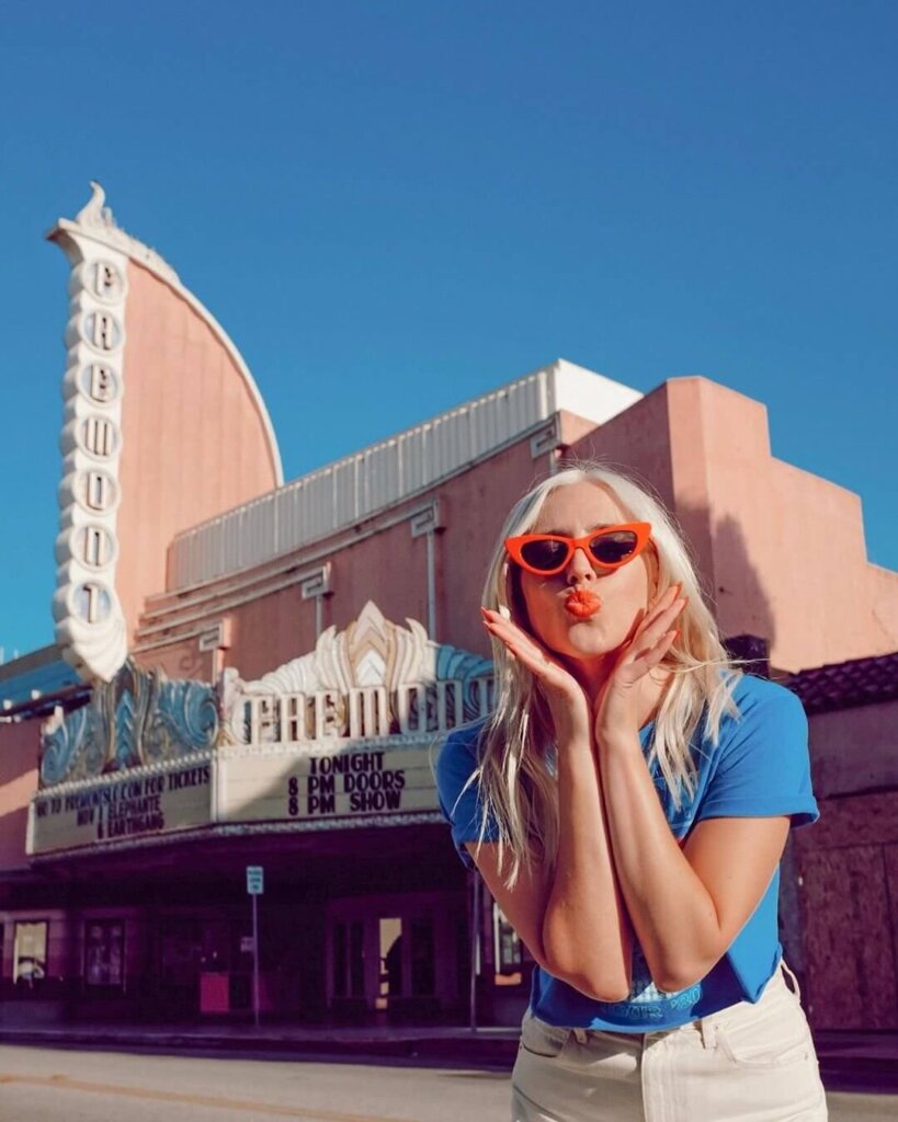 A young blond-haired woman with red, retro sunglasses blowing a kiss in front of the light-pink Fremont Theater in San Luis Obispo.