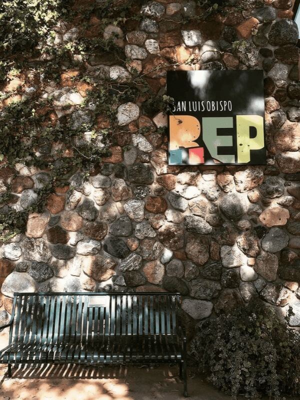 A bench in front of the cobble-stone wall of the SLO Rep Theater, with a colorful sign on the wall. Photo by @astardanced4me