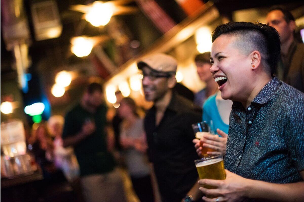 Laughing people in a dim bar, holding pints of beer in hand.