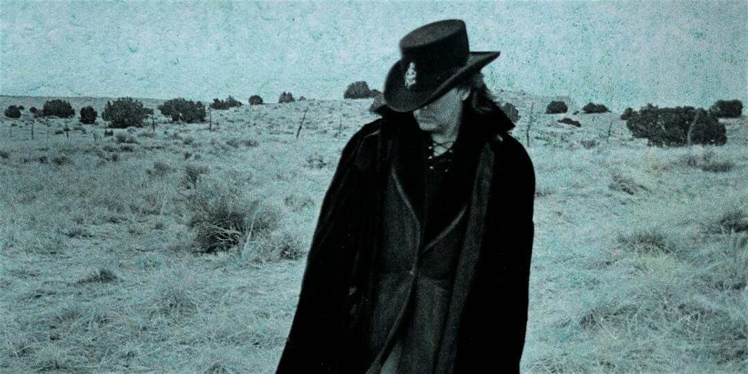 Marty Stuart in a trench coat and cowboy hat in a blurry desert.
