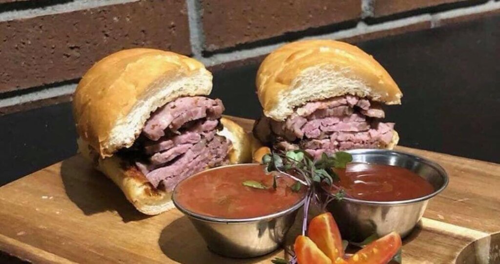 2 halves of a tri-tip sandwich with sauce in front of a brick wall in the Rib Line restaurant.